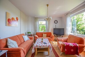 Olohuoneessa on oranssit sohvat, kukkapintainen pöytäliina päällystetty sohvapöytä, televisio ja suuri ikkuna, josta avautuu näkymä vehreään maisemaan.