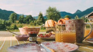 Una mesa de desayuno con pan, huevos y una jarrón de jugo de naranja, con montañas al fondo.