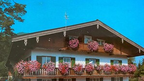 Un edificio de dos pisos con balcones decorados con flores, techo inclinado y antena visible.