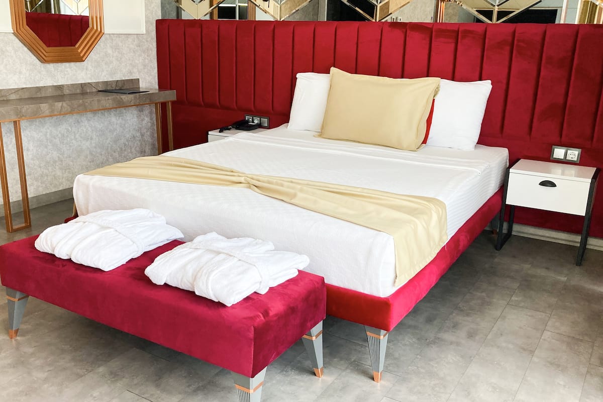 A hotel room with a large bed, a bedside table, a red headboard, and a red bench with towels.