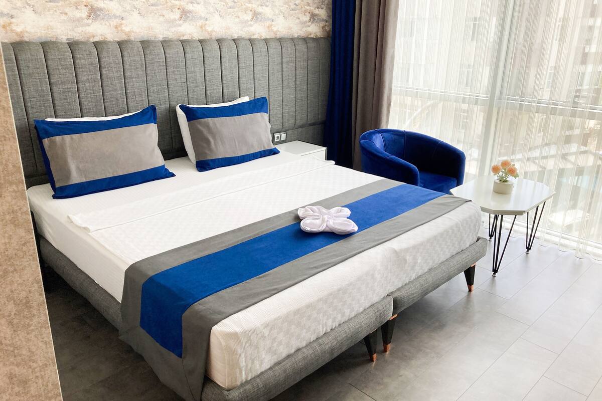 A hotel room with a large bed, a headboard, two pillows, a blue chair, a small table with a vase, and a view through sheer curtains.