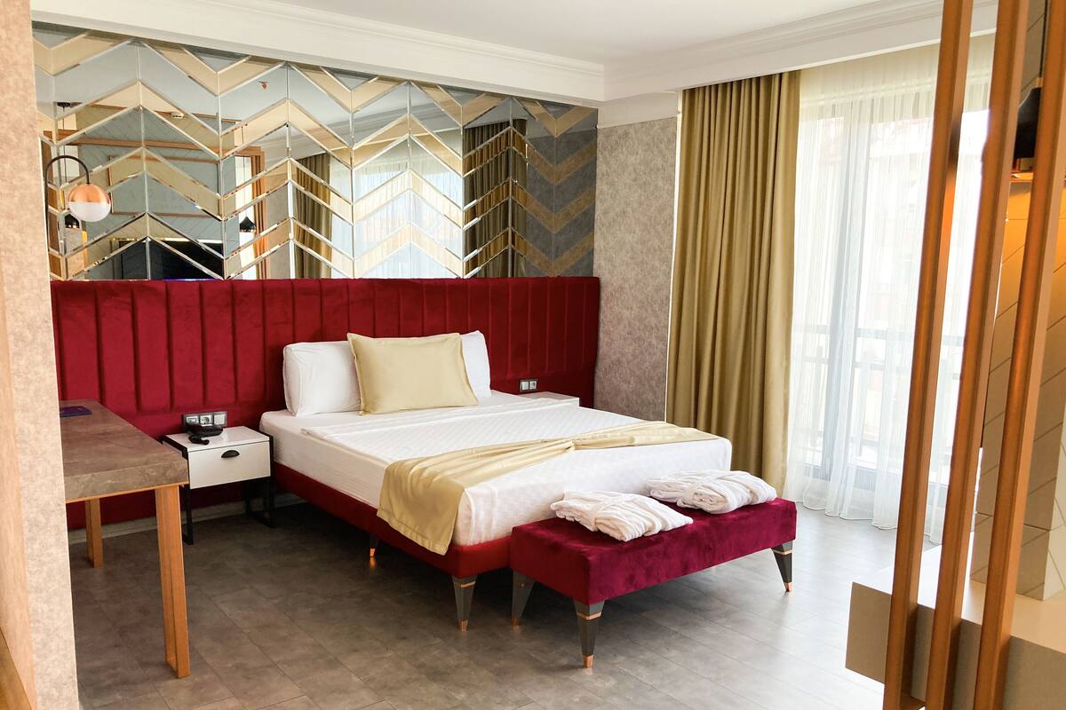 A hotel room with a red upholstered headboard, a bed with white linens, a bedside table with a lamp, and a large window with sheer curtains.