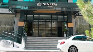 Un moderno edificio de hotel con una fachada azul turquesa destacada, una entrada visible con escaleras y un coche blanco estacionado al frente.