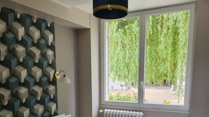 A bedroom with a bed, a wall-mounted lamp, a window with a view of greenery, and a radiator.