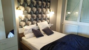 A bedroom with a bed, pillows, a bedside table, a dresser, a wall with geometric patterns, and a window with a view.