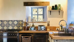 Een keuken met een fornuis, toaster en diverse keukengerei. Er is een raam met uitzicht op een stenen muur, en planken met potten.