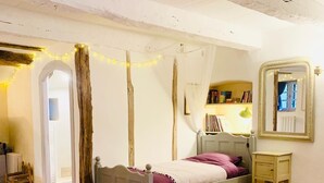 Een slaapkamer met een wit bed, een nachtkastje, een spiegel en een boekenplank.