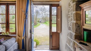 A room with a large open door leading to a garden, a wooden cabinet, and a window with curtains. - Gîte du Château de Montafilan (Corseul)