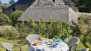 A rustic stone building with a sloped roof and a small window. In front, there is a round table with a white tablecloth and four checkered chairs. - Gîte du Château de Montafilan (Corseul)