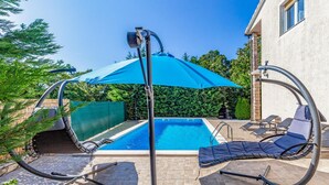 En swimmingpool med blå parasol, to liggestole og en pergola.