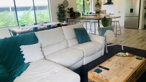 A modern living room with a white sofa, teal cushions, a wooden coffee table, and a kitchen area in the background. - Modern country house set in the heart of the vines (McLaren Flat)