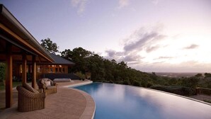 Una piscina infinita moderna con vista al horizonte de la ciudad, rodeada de exuberante vegetación y un pabellón de madera con mobiliario de mimbre.