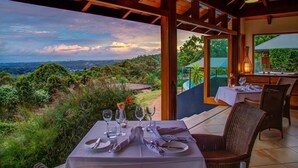 Un comedor con una mesa dispuesta para cuatro personas, con vistas a un paisaje exuberante y una silueta de colinas y el horizonte de una ciudad.