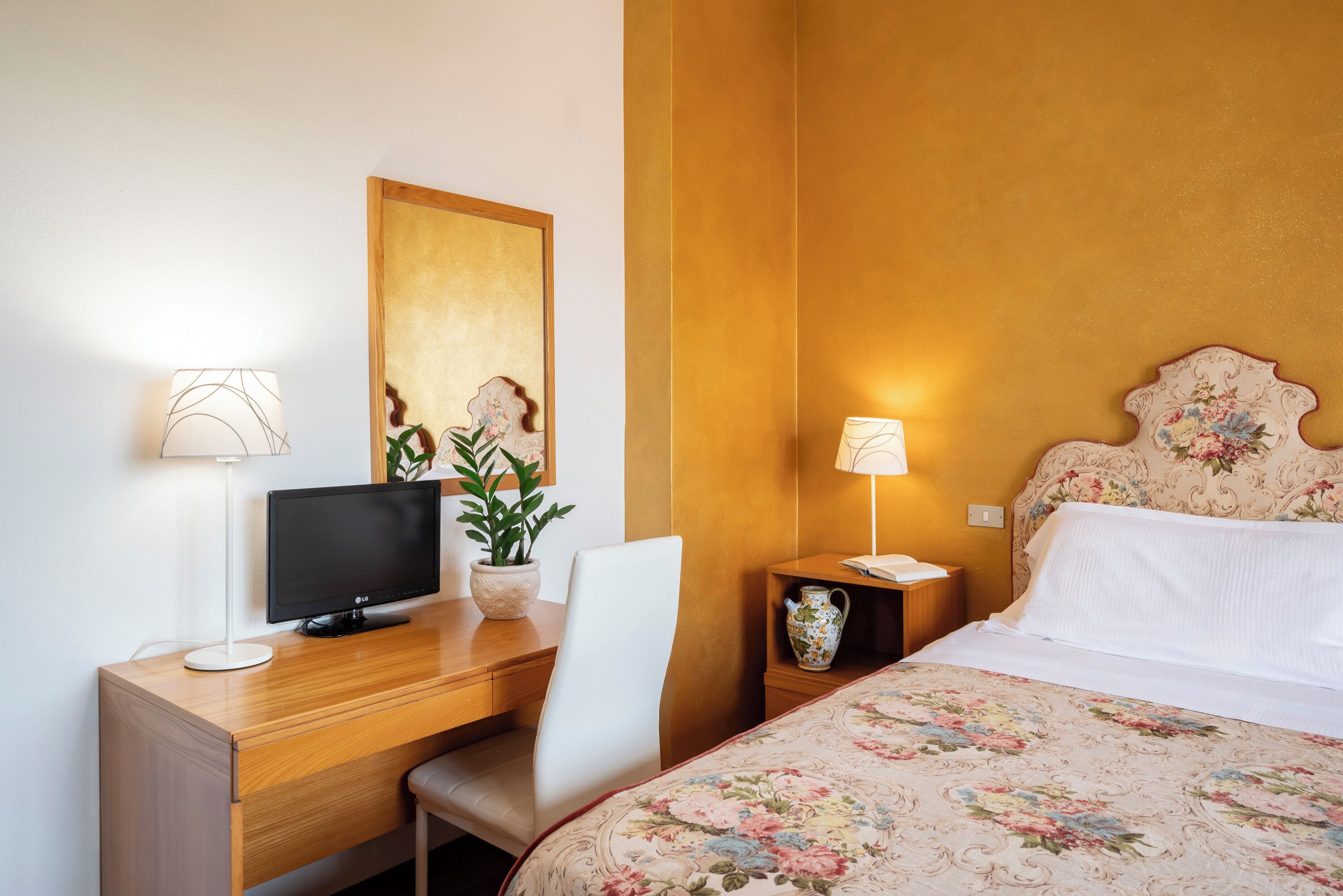 Une chambre d’hôtel avec un bureau en bois, une télévision, une plante en pot, une table de chevet avec une lampe et un lit recouvert d’une courtepointe à motifs floraux.
