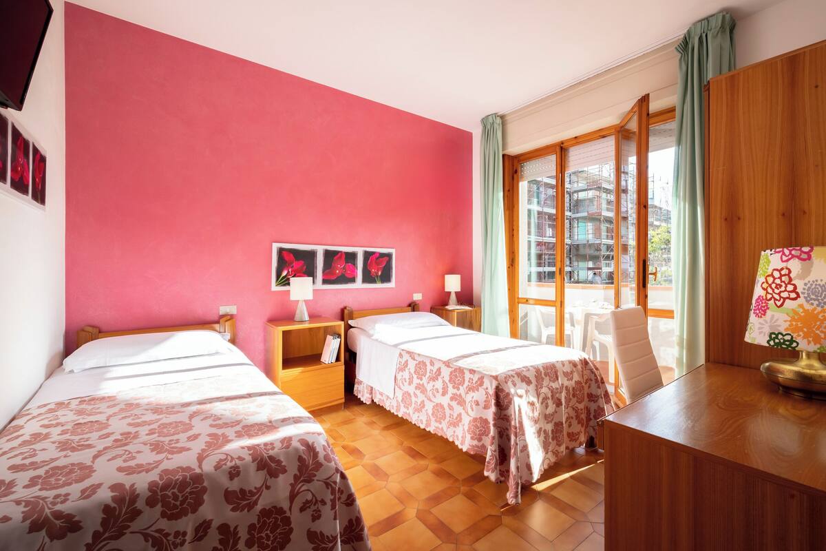 A hotel room with two beds, a desk, a chair, and a large window with a view of buildings.