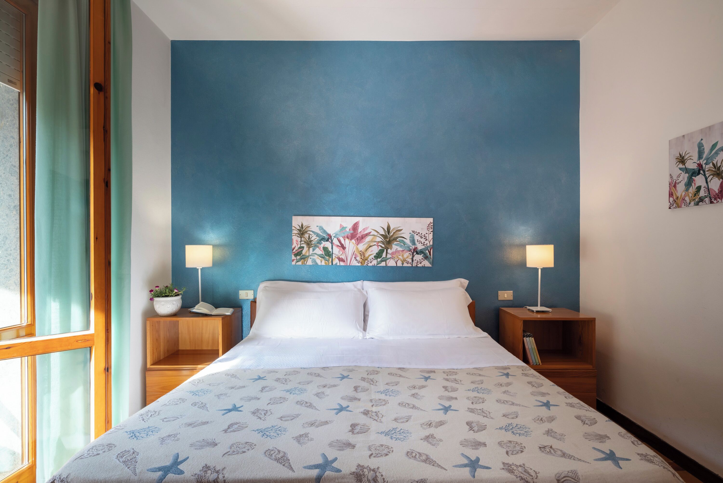 Une chambre avec un mur d’accent bleu, un lit recouvert d’une couvre-lit à motifs d’étoiles de mer, deux tables de chevet avec des lampes et une fenêtre avec des rideaux.