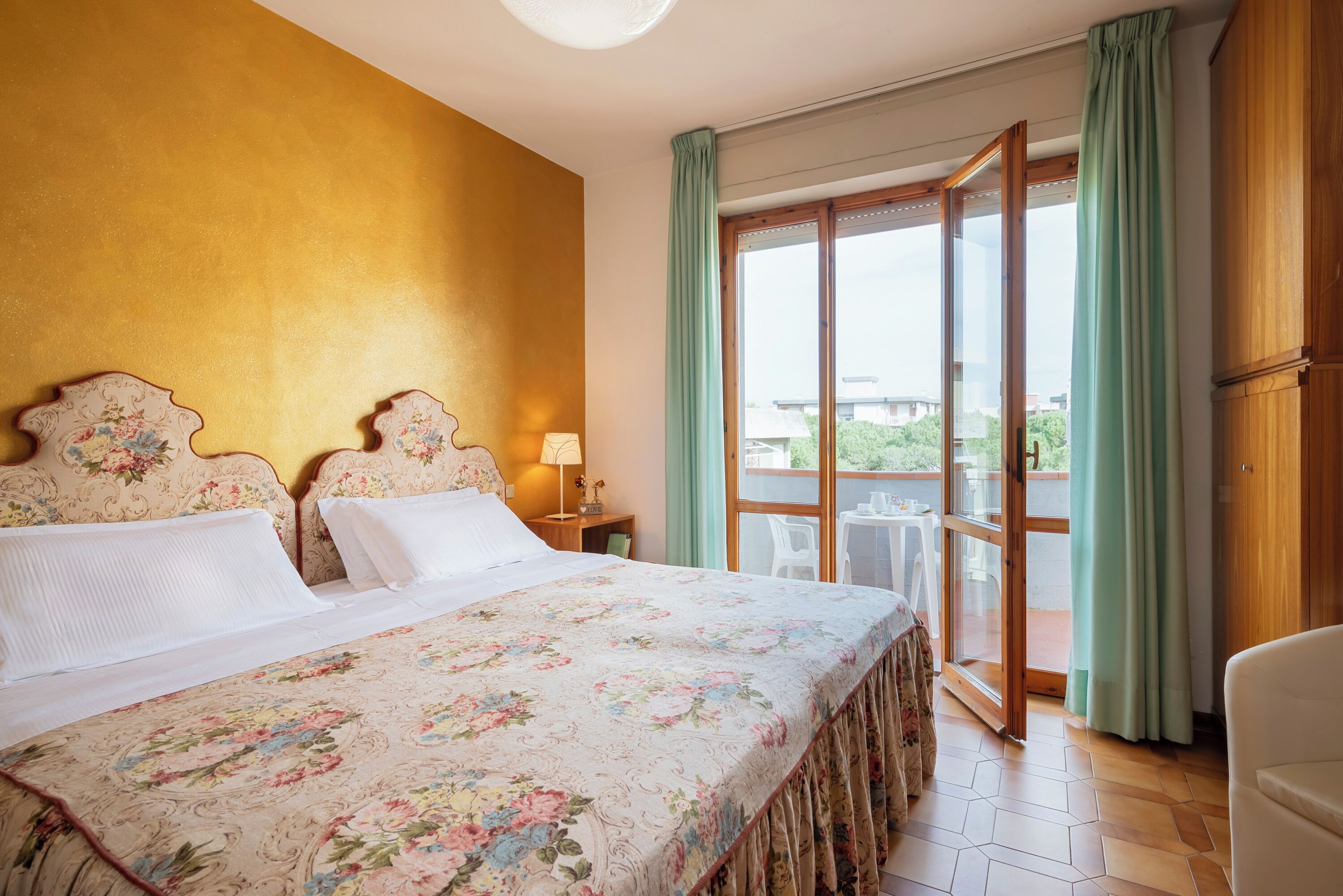 A hotel room with a bed, a nightstand, a lamp, a balcony with a table and chairs, and a view of the outside.