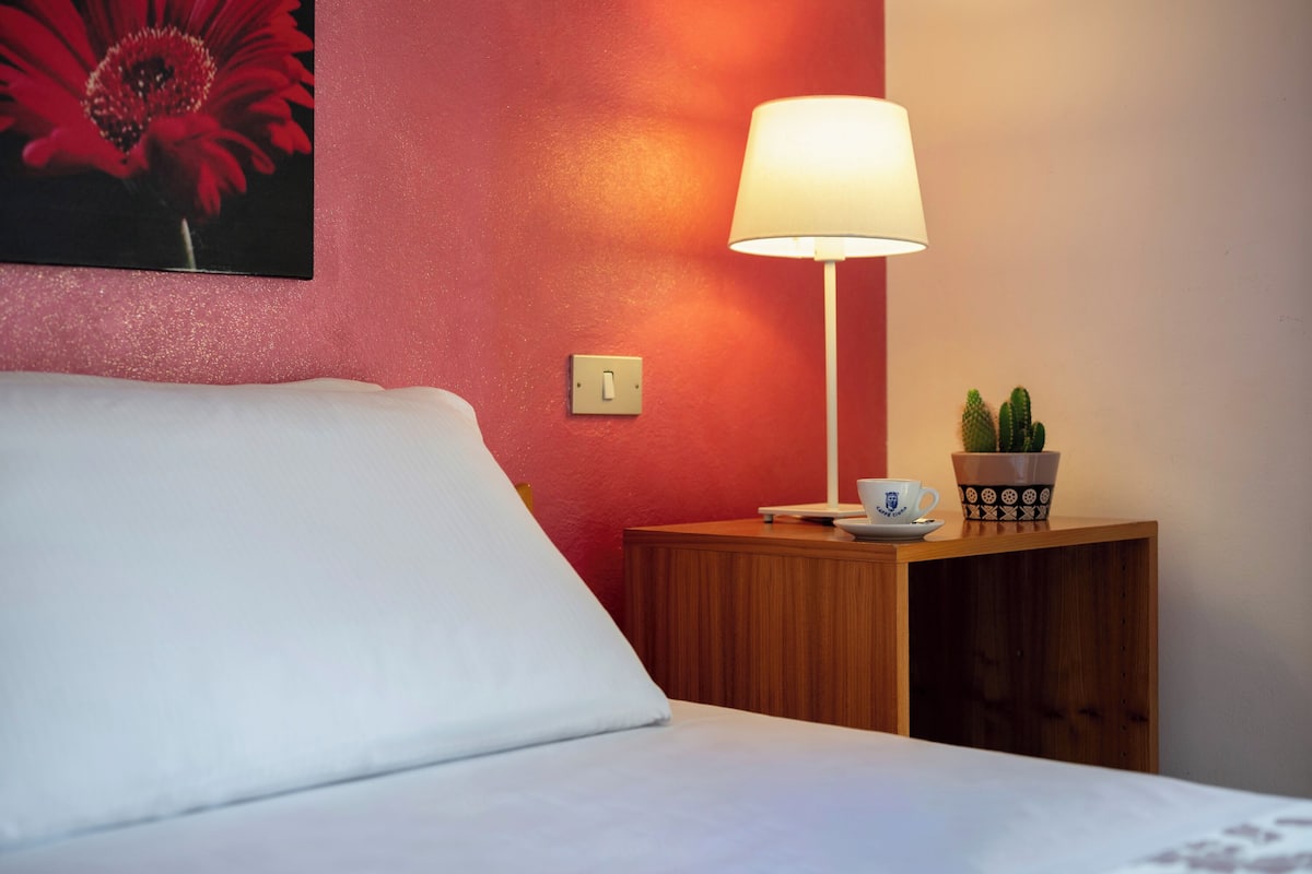 A hotel room with a bed, a nightstand with a lamp, a potted plant, and a painting on the wall.