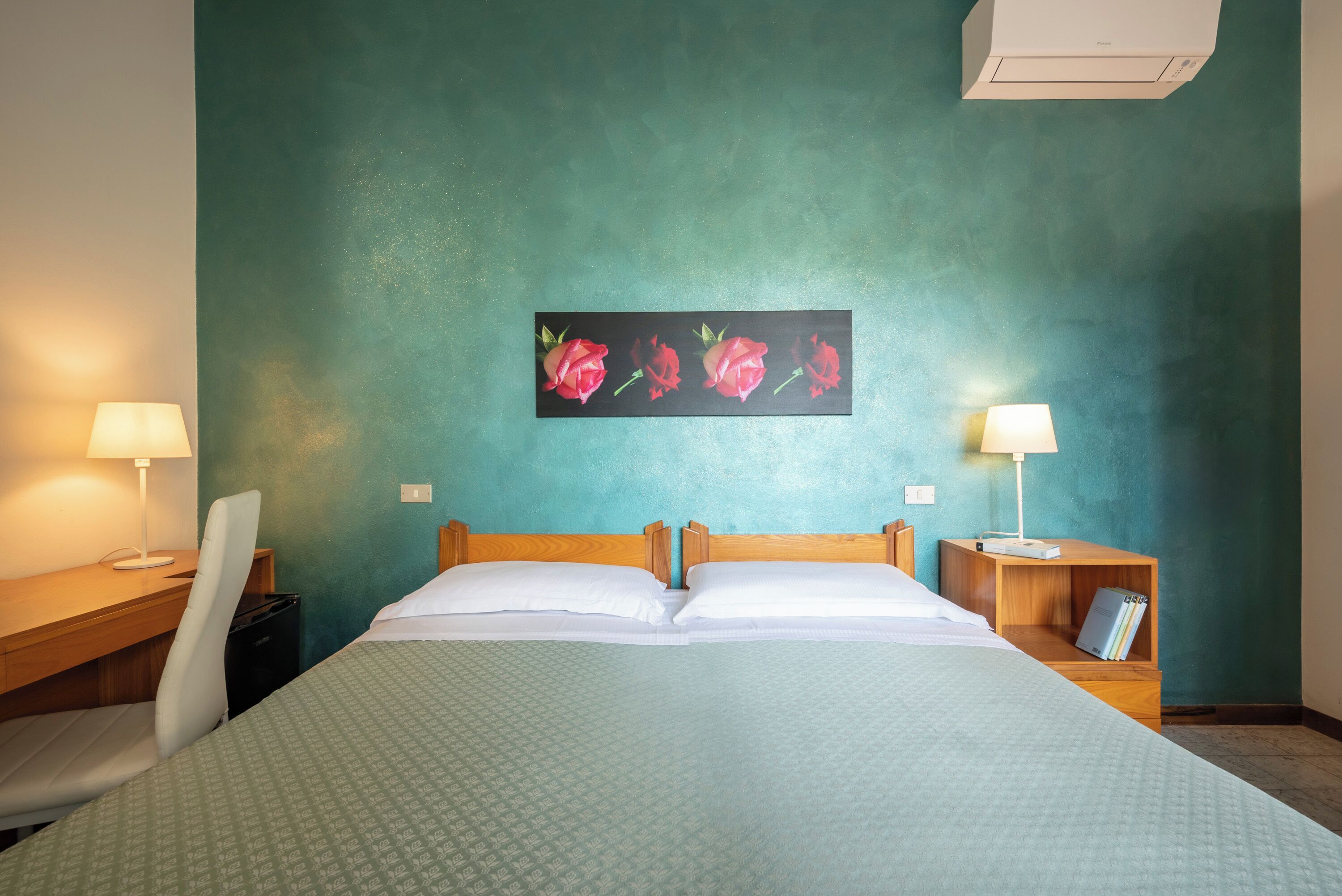 A hotel room with a bed, two bedside tables with lamps, a desk, a chair, and a painting of roses on the wall.