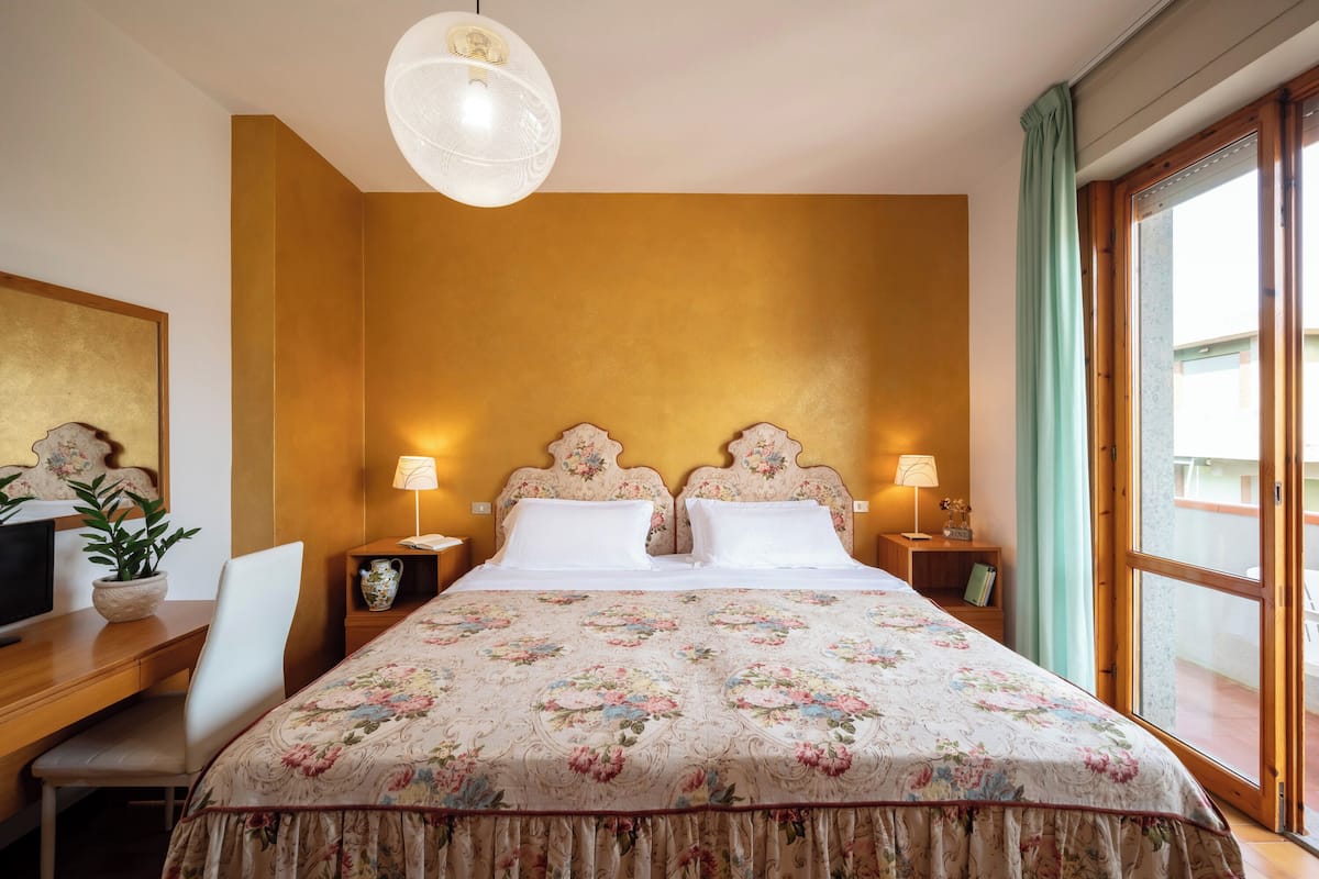 A hotel room with a large bed, two bedside tables with lamps, a desk with a chair, a potted plant, and a view of the outside through a large window.