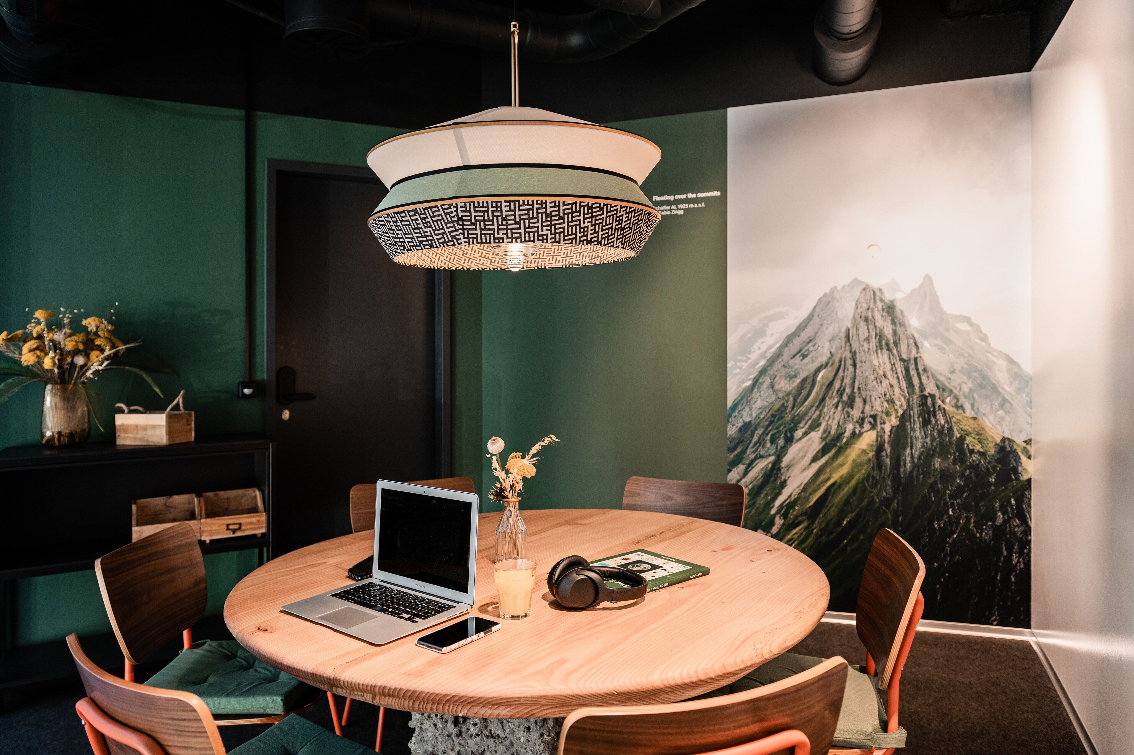 Een ronde houten tafel met een laptop, een glas melk, een koptelefoon en een vaas met bloemen.