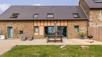 Een gebouw van steen met een schuin dak, een terras met een tafel en stoelen, en een barbecue.