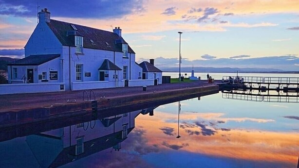 A white building with a sloped roof and a dock extending into a calm body of water. - 2 Bedroom House  with the Caledonian Canal at rear. (Inverness)