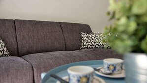 A grey sofa with patterned cushions, a blue table with two cups and saucers, and a green plant in the foreground. - Apt. Neuf à 50m du Port - Parking Privé, Ascenseur (Saint-Valery-sur-Somme)
