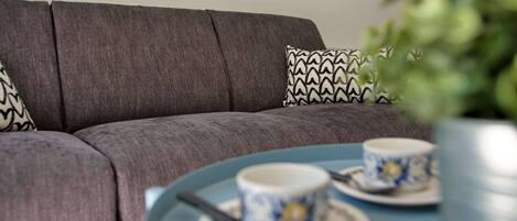 A grey sofa with patterned cushions, a blue table with two cups and saucers, and a green plant in the foreground.