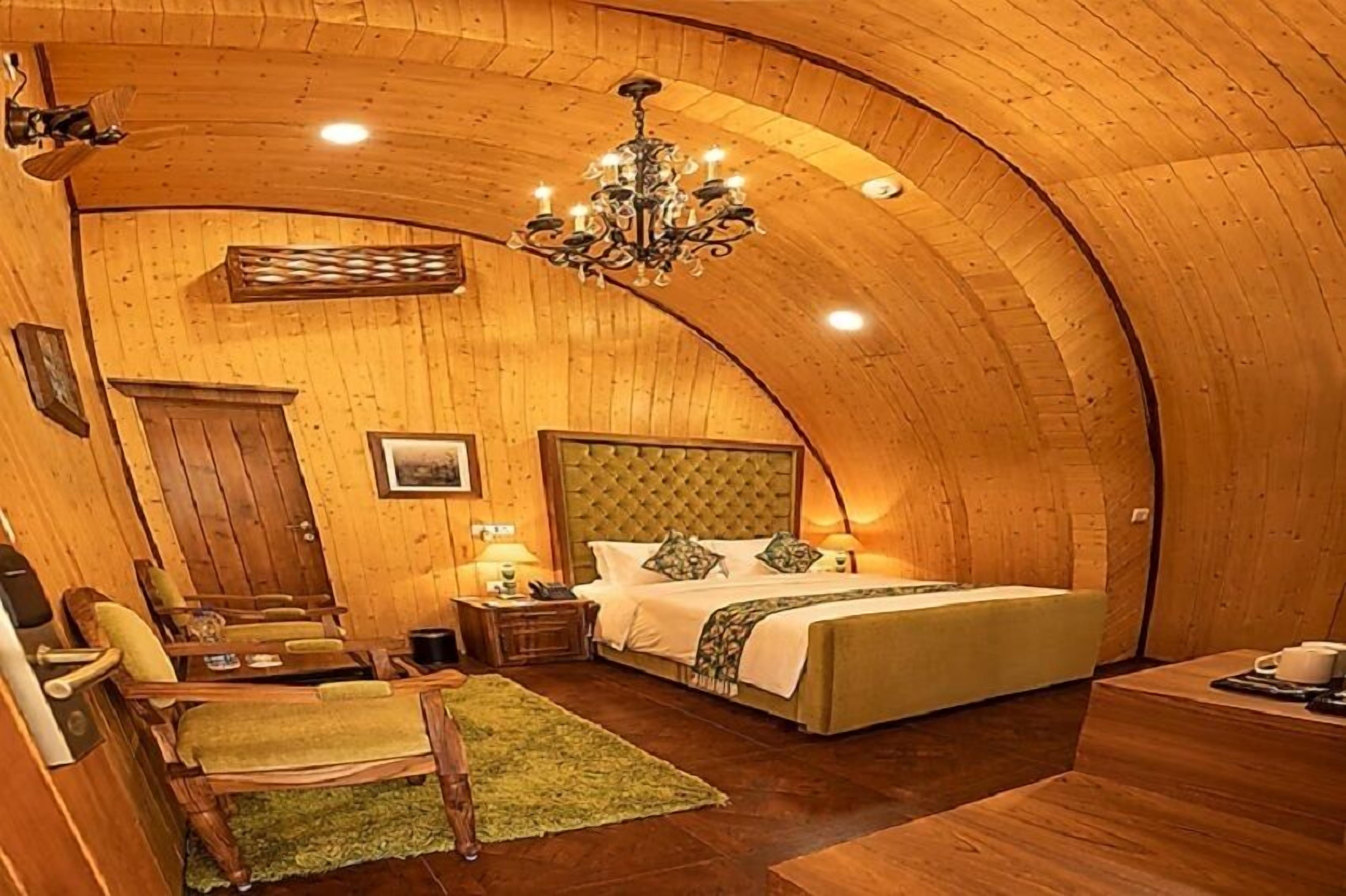 A cozy room with a curved wooden ceiling, a bed with a tufted headboard, a chandelier, and a small table with a chair.