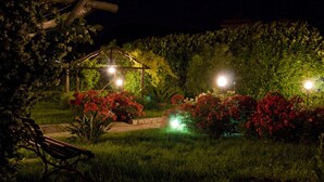 Ein Garten bei Nacht mit einer Bank, beleuchtet durch Gartenleuchten, umgeben von üppigem Grün und blühenden Pflanzen.