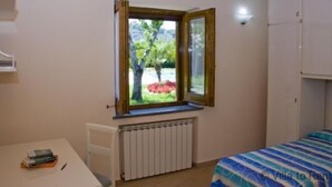 Ein Schlafzimmer mit Fenster, Heizkörper, Bett und Tisch mit Büchern.