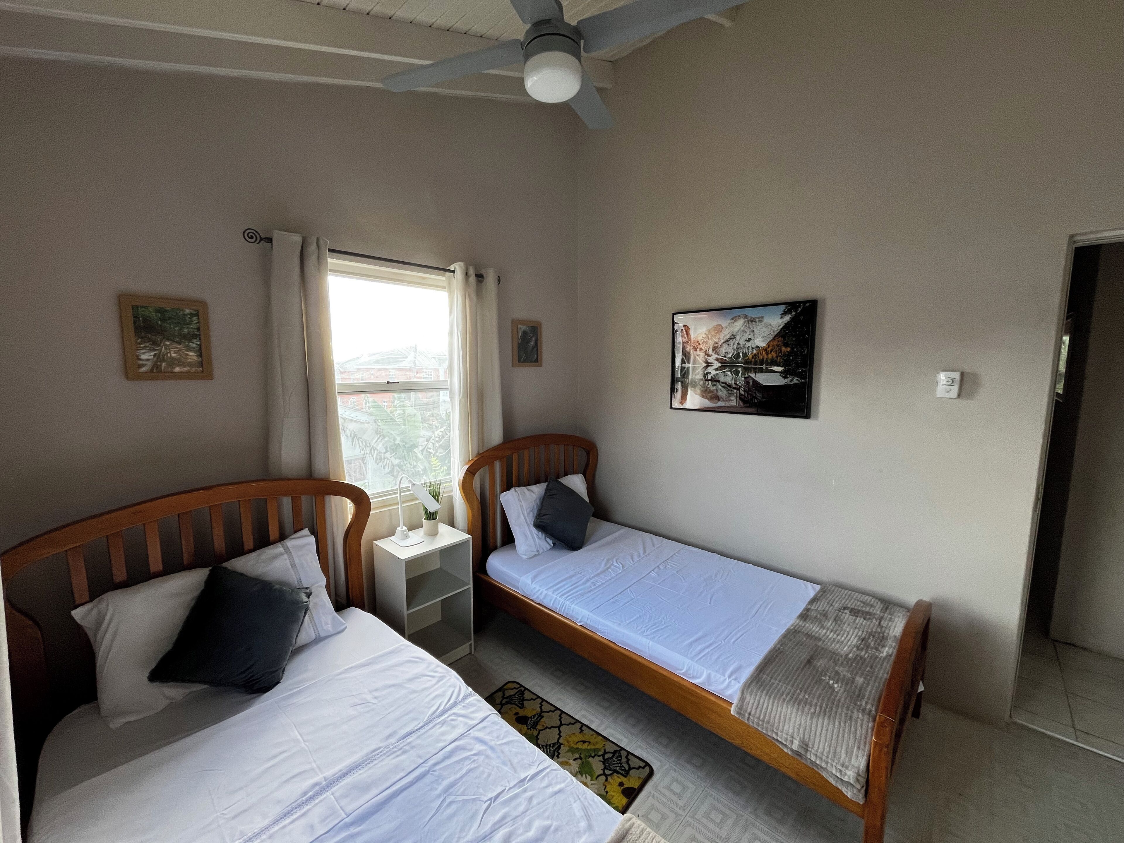 Een kamer met twee éénpersoonsbedden, een nachtkastje met lamp, een plafondventilator, een raam met gordijnen en een ingelijste foto aan de muur.