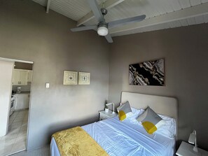 Een slaapkamer met een bed, een plafondventilator, een nachtkastje, een lamp en een schilderij aan de muur.