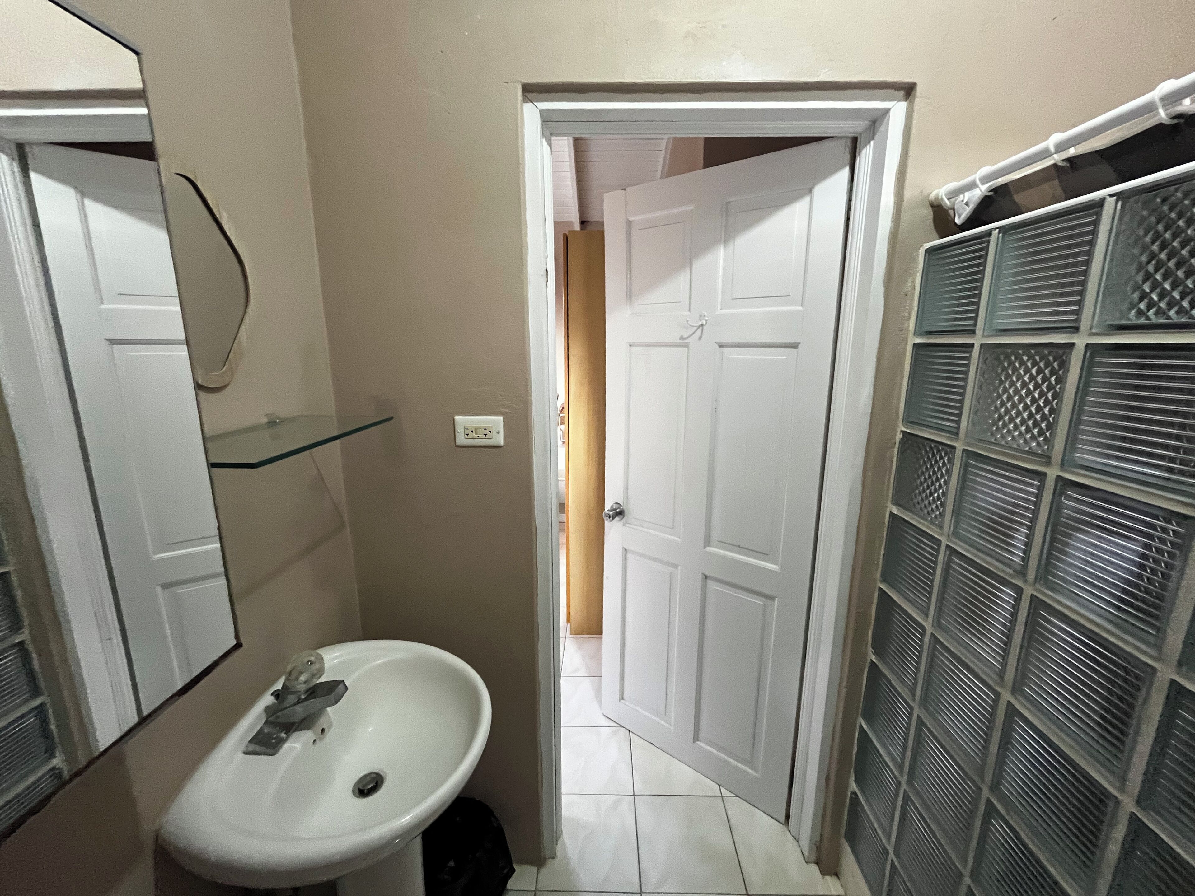 Een smalle badkamer met een wit wastafel, een spiegel en een deur naar een andere kamer.