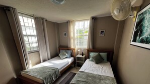 Una habitación con dos camas individuales, un ventilador de pie, una mesita de noche con lámpara y una ventana con cortinas.