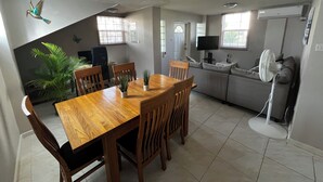 Un comedor con mesa y sillas de madera, una cocina con televisión y ventilador, y un ventanal con persianas.