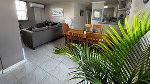 Un salón con sofá, mesa de comedor, sillas y un ventilador. Se observa una zona de cocina con armarios y un refrigerador al fondo.