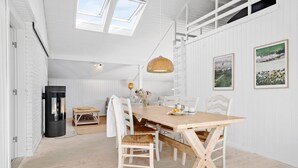 Un coin repas avec une table en bois et des chaises blanches, un poêle noir et deux tableaux représentant des paysages, encadrés et accrochés au mur.