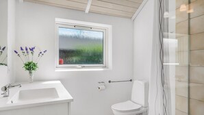 Une salle de bain avec un lavabo blanc, des toilettes, une fenêtre donnant sur un espace verdoyant et un espace douche.