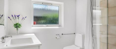 A bathroom with a white sink, a toilet, a window with a view of greenery, and a shower area.