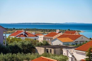 A coastal village with terracotta-roofed houses, a clear blue sea, and distant mountains.