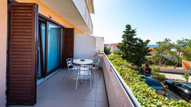 A balcony with a table and chairs, a sliding glass door, and a view of the sea and greenery. - Apartments 4 You (Novalja)