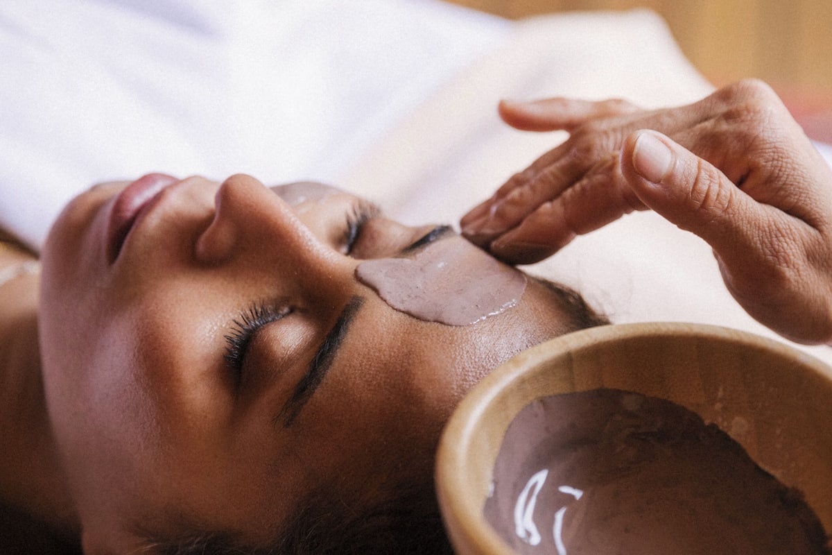 Una persona recostada con un recipiente de líquido marrón sobre el rostro, probablemente un tratamiento facial.