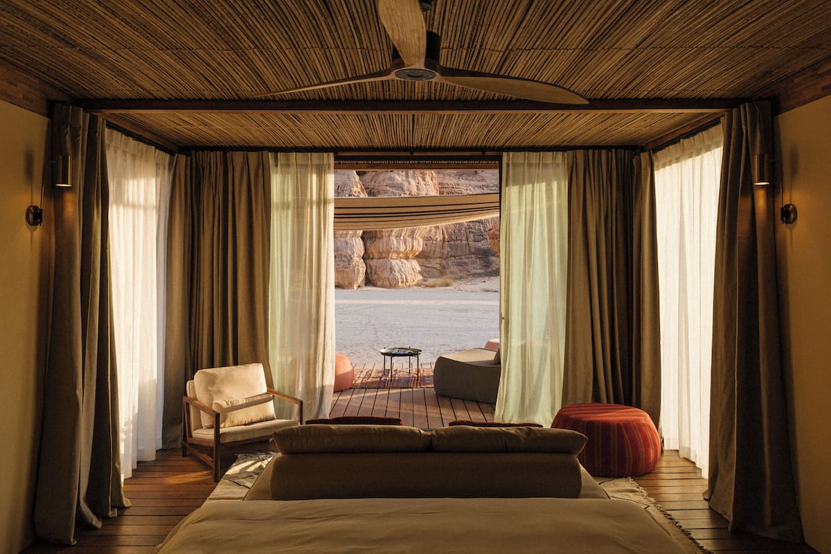 Un dormitorio con una cama grande, una silla, una mesita y vistas a un paisaje rocoso a través de cortinas translúcidas.