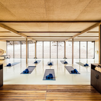 A spacious room with a wooden floor, large windows, and a view of a desert landscape. There are several blue mats arranged in the center, and wooden cabinets along the walls.