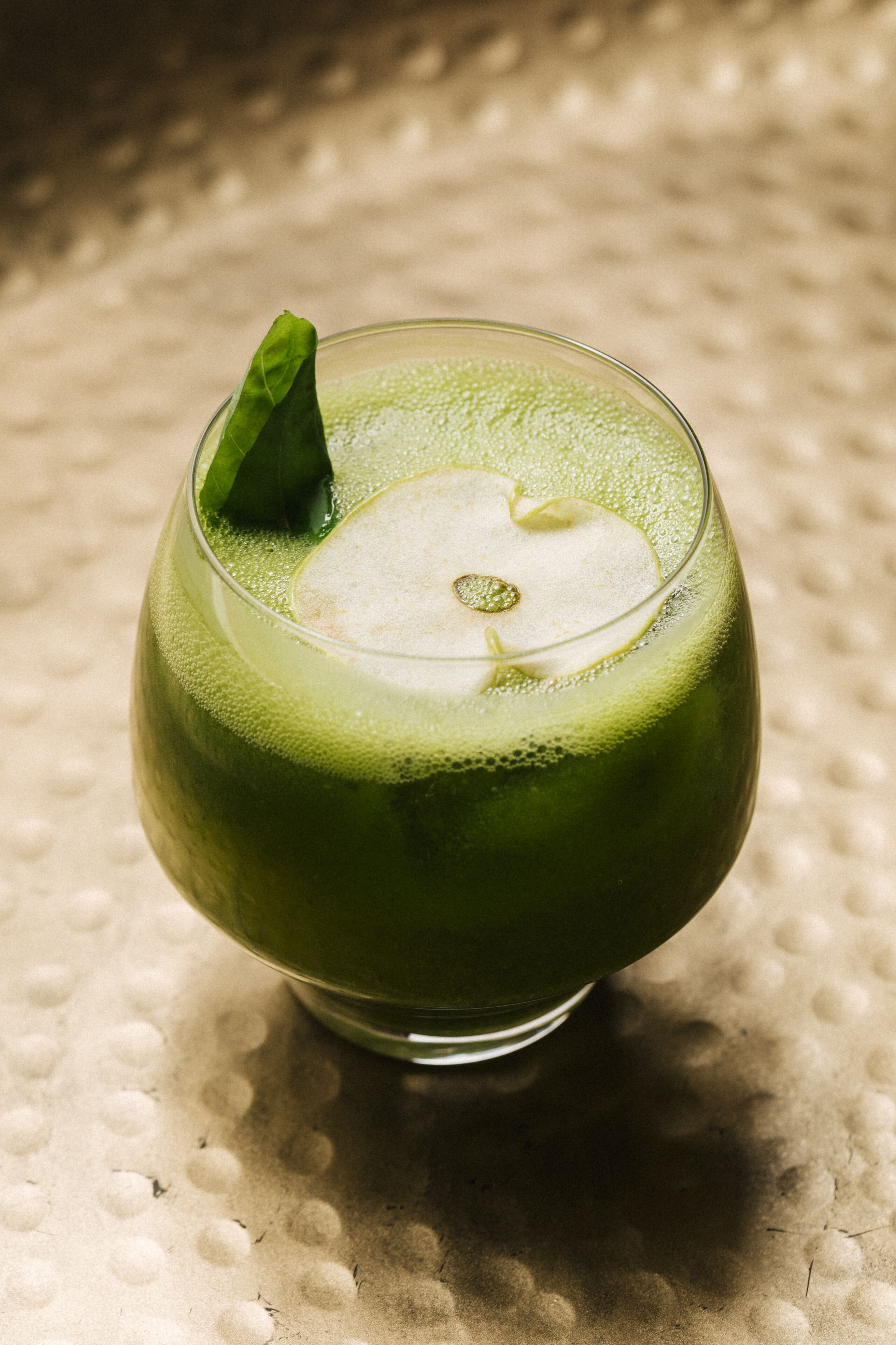 Ein Glas grünes Getränk mit Apfelscheibe und einem Blatt als Dekoration auf einer strukturierten Oberfläche.