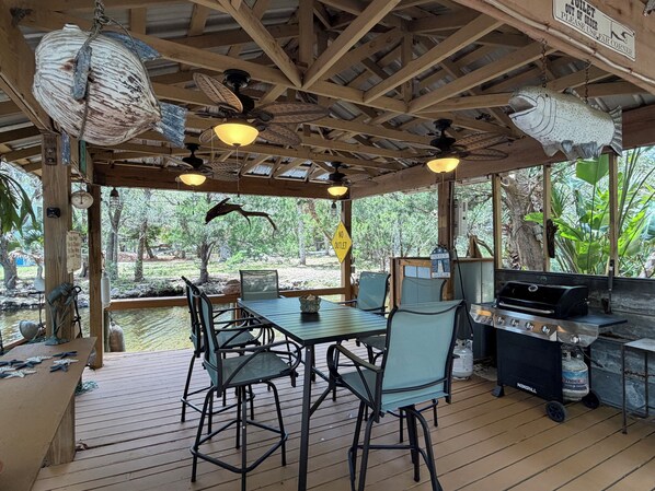 A covered outdoor dining area with a grill, wooden ceiling, and natural surroundings.