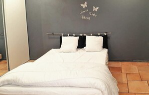 Un lit bien fait, avec une literie blanche et deux oreillers, une tête de lit sombre et un mur décoré de motifs de papillons, sur lequel on peut lire les mots « Amour, Petit, Bagu ».