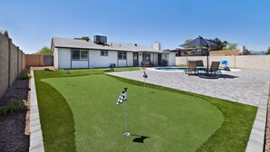 Un patio trasero con césped artificial, una piscina y un área de terraza con sillas y una mesa.